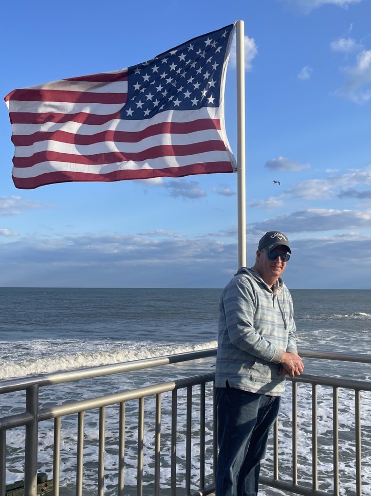 Steve DeVoll standing by the ocean with an American flag in the background